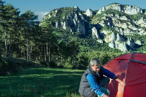 Actor portrayal of a man setting up a tent outdoors. Behind him, a forest leads in to a mountain range
